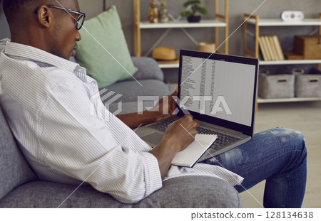 African American business man working on a laptop with tables making notes sitting on sofa. African American business man working on a laptop with tables making notes sitting on sofa. 128134638