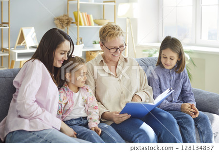 Happy family day at home, granny older woman, mother, two kids sitting on sofa reading book together 128134837