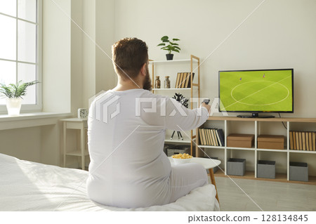 Funny lazy fat man in pyjamas sitting on bed at home and watching football on TV 128134845