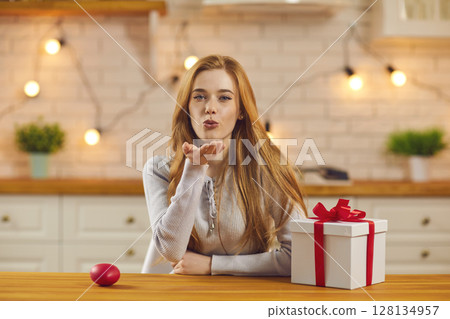 Pretty young woman sitting at kitchen table with Valentine present and blowing a kiss at camera 128134957