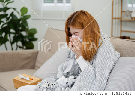 Young Woman Suffering from a Cold, Wrapped in a Blanket, Sneezing into a Tissue on a Sofa at Home Young Woman Suffering from a Cold, Wrapped in a Blanket, Sneezing into a Tissue on a Sofa at Home 128135455