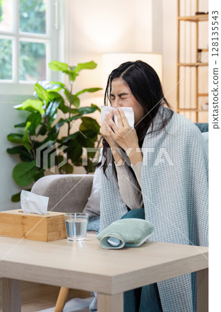 Woman Sneezing into Tissue While Sitting on Couch, Wrapped in Blanket, Battling Cold in Cozy Living Room Woman Sneezing into Tissue While Sitting on Couch, Wrapped in Blanket, Battling Cold in Cozy Living Room 128135543