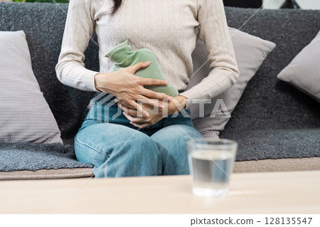 Woman Using Hot Water Bottle for period pain relief on sofa. sick at home concept 128135547