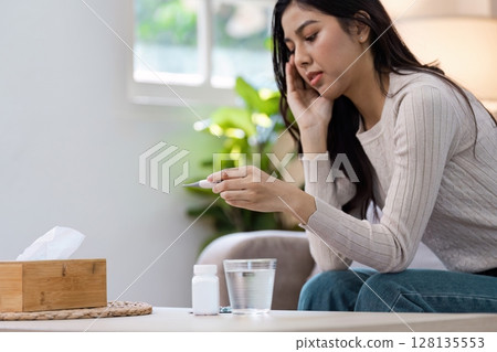Woman Checking Thermometer While Experiencing Fever and Headache at Home, Sitting on Sofa with Medicine and Tissue Box Woman Checking Thermometer While Experiencing Fever and Headache at Home, Sitting on Sofa with Medicine and Tissue Box 128135553