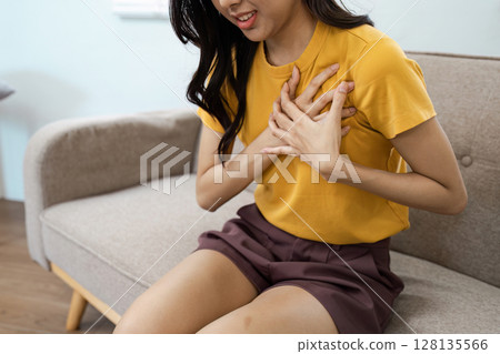 Woman Experiencing Chest Pain While Sitting on Sofa, Concept of Heart Attack, Health Emergency, and Cardiovascular Issues 128135566