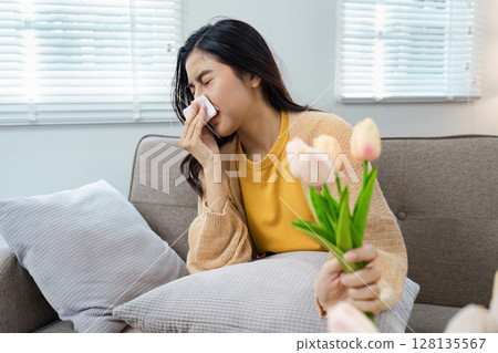 Young Woman Sneezing Due to Pollen Allergy While Holding Tulips in a Bright Living Room 128135567