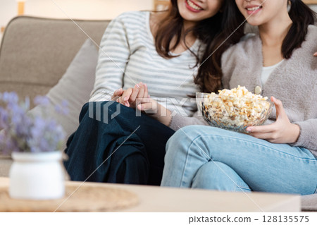 Cozy LGBT Couple Enjoying Quality Time Together at Home, Relaxing on the Couch, Sharing a Bowl of Popcorn, and Holding Hands in a Warm and Comfortable Living Room Setting Cozy LGBT Couple Enjoying Quality Time Together at Home, Relaxing on the Couch, Sharing a Bowl of Popcorn, and Holding Hands in a Warm and Comfortable Living Room Setting 128135575