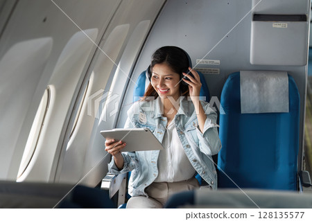 Young Woman Enjoying In-Flight Entertainment on a Modern Airplane, Smiling and Relaxed While Using a Tablet and Wearing Headphones, Capturing the Joy of Air Travel and the Comfort of Modern Aviation 128135577