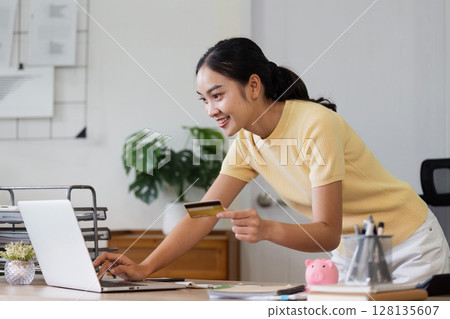 Young Woman Using Credit Card for Online Shopping on Laptop at Home Office, Smiling and Excited, Modern E-commerce Concept 128135607