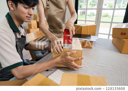 Small Business owner Packing Boxes for Shipment in a Modern Home Office with Natural Light and Large Windows 128135608
