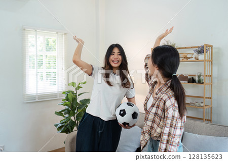 Excited LGBT Lesbian Couple Cheering for Sports Team at Home, Celebrating Victory with Joyful Expressions, Holding Soccer Ball in Modern Living Room Excited LGBT Lesbian Couple Cheering for Sports Team at Home, Celebrating Victory with Joyful Expressions, Holding Soccer Ball in Modern Living Room 128135623