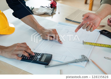 Architect, Engineers Collaborating on Blueprint Design with Tools and Safety Helmet on Desk, Discussing Construction Plans and Measurements in Modern Office Environment 128135628
