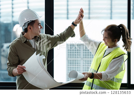 Two Engineers Collaborating on a Project, Reviewing blueprint and Celebrating Success in a Modern Office Setting with Natural Light 128135633