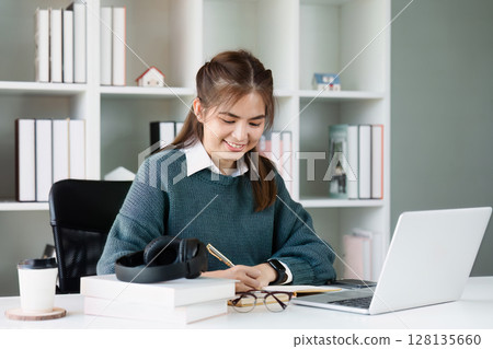 Young Woman Managing Finances at Home, Smiling While Reviewing Bills and Saving Plans on Laptop, Cozy Home Office Setting 128135660