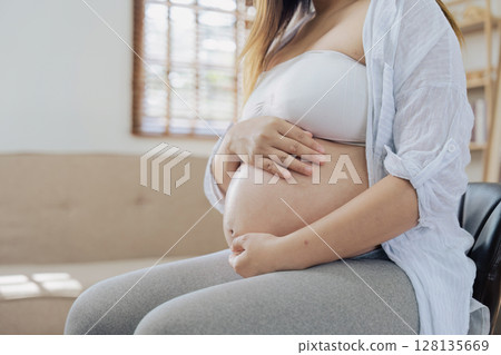 pregnancy Woman Sitting on Sofa Holding Belly, Expecting Mother in Comfortable Home Setting, Maternity and Parenthood Concept, Close Up of Baby Bump, Anticipation and Preparation for New Life 128135669