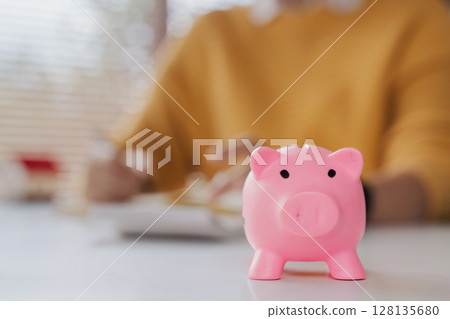 Close up of Pink Piggy Bank with woman Calculating Bills in Background, Concept of Saving Money and Financial Planning 128135680