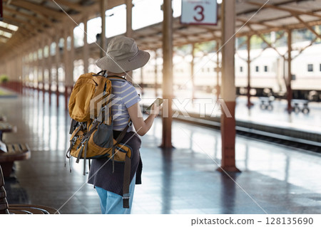 Solo Traveler with Backpack and Hat Waiting at Train Station Platform, Checking Phone for Travel Updates, Adventure and Exploration Concept Solo Traveler with Backpack and Hat Waiting at Train Station Platform, Checking Phone for Travel Updates, Adventure and Exploration Concept 128135690