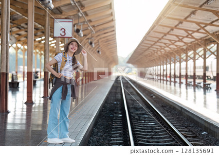 Young Female Traveler at Train Station Platform Ready for Adventure, Wearing Backpack and Hat, Smiling and Looking Excited for Journey, Sunlight Streaming Through Wooden Roof Structure Young Female Traveler at Train Station Platform Ready for Adventure, Wearing Backpack and Hat, Smiling and Looking Excited for Journey, Sunlight Streaming Through Wooden Roof Structure 128135691