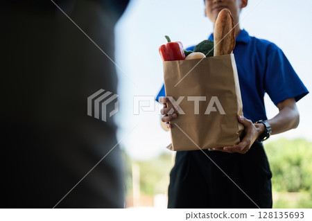 Delivery Service Concept - Close-Up of a Delivery Person Handing Over a Paper Bag of Groceries to a Customer at the Doorstep, Representing Modern Convenience and Efficient Service 128135693
