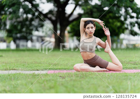 Young Woman Practicing Yoga Outdoors in a Park, Demonstrating Flexibility and Balance in a Natural Setting, Promoting Health and Wellness Through Mindful Exercise and Meditation 128135724
