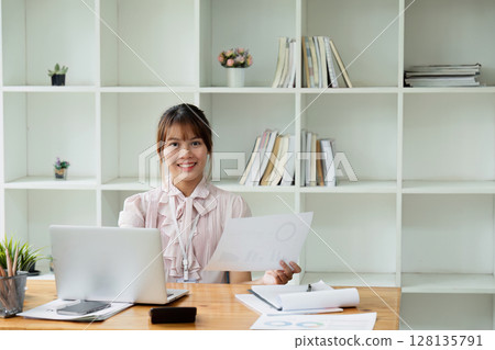 Young Business Professional Woman Working at Desk in Modern Office with Laptop and Documents, Smiling Confidently, Surrounded by Bookshelves and Office Supplies Young Business Professional Woman Working at Desk in Modern Office with Laptop and Documents, Smiling Confidently, Surrounded by Bookshelves and Office Supplies 128135791