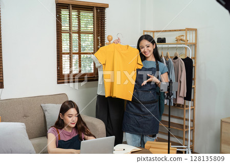 Young Online E-Business Owners Working from Home, Showcasing Clothing Products, and Engaging with Customers via Live Streaming and Laptop in a Modern Home Office Setting 128135809