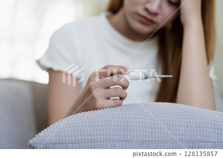 Young Woman Feeling Unwell at Home Holding Thermometer, Resting on Couch, Battling Illness, Experiencing Fever and Fatigue, Indoor Scene, Healthcare and Sickness Concept 128135857