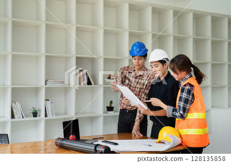 Team of Engineers and Architects Collaborating on Construction Project in Modern Office with Shelves in Background Team of Engineers and Architects Collaborating on Construction Project in Modern Office with Shelves in Background 128135858