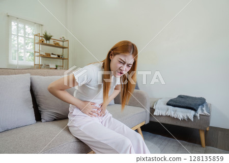 Young Woman Feeling Unwell at Home, Sitting on Couch with Stomach Pain, Indoor Scene Depicting Sickness, Discomfort, and Illness in a Modern Living Room Setting 128135859