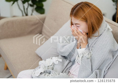 Woman Sick at Home Wrapped in Blanket Sneezing into Tissue on Couch in Living Room - Concept of Illness, Cold, Flu, and Resting at Home 128135878