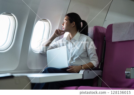 Business Travel. Young woman working on laptop while gazing out airplane window. Business Travel. Young woman working on laptop while gazing out airplane window. 128136015