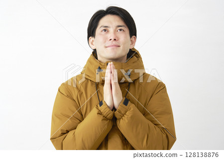 A man wearing a down jacket praying with his hands together on a white background 128138876