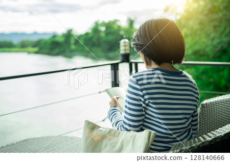 Woman sitting on chair reading a book on balcony overlooking river during golden hour morning. Self care, digital detox, and mindful slow living. Emotional wellness in riverside retreat. Quiet luxury. 128140666