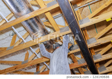 Construction worker wearing protective gear installs spray foam thermal barrier on wooden beams in ceiling. Construction worker wearing protective gear installs spray foam thermal barrier on wooden beams in ceiling. 128140686