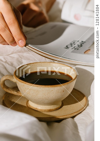 Cozy morning scene with hand flipping magazine pages next to rustic ceramic cup of black coffee  128141884