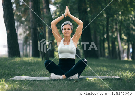 Senior Woman in Sukhasana Yoga Pose Practicing Mindfulness with Raised Hands Outdoors 128145749