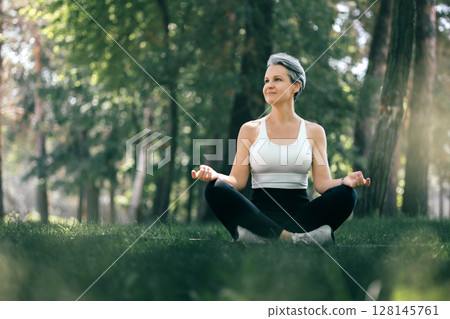 Smiling Mature Woman Meditating Outdoors in Sukhasana Yoga Pose Surrounded by Nature Smiling Mature Woman Meditating Outdoors in Sukhasana Yoga Pose Surrounded by Nature 128145761