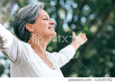 Mature Woman Smiling with Open Arms and Closed Eyes Enjoying Nature in a Peaceful Forest 128145762