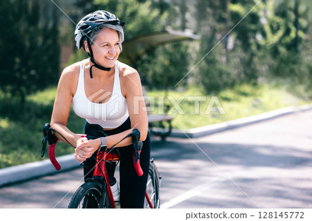 Confident senior woman cyclist smiling while resting with road bike on sunny path in summer Confident senior woman cyclist smiling while resting with road bike on sunny path in summer 128145772