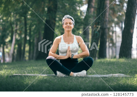 Mature Woman Meditating with Hands in Namaste Pose Outdoors in a Peaceful Forest Setting 128145773