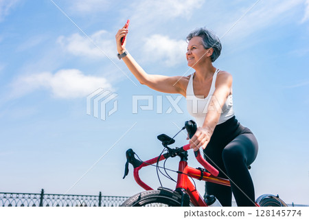 Senior Woman Taking Selfie on Smartphone While Riding Bicycle Along Seafront on Sunny Day Senior Woman Taking Selfie on Smartphone While Riding Bicycle Along Seafront on Sunny Day 128145774