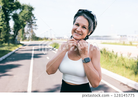 Confident woman fastening helmet before bike ride on a sunny summer day in city park lane 128145775
