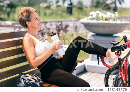 Sporty woman drinks water while resting on a bench with bicycle on a sunny day in the park 128145776