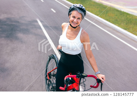 Smiling Mature Woman Standing with Bicycle on Urban Road in Helmet and Activewear Smiling Mature Woman Standing with Bicycle on Urban Road in Helmet and Activewear 128145777