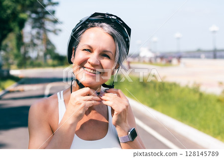 Smiling active woman fastening bicycle helmet before cycling on a sunny outdoor trail 128145789