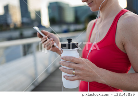 Runner checking her performance on a smartphone during her morning run across a city bridge. Runner checking her performance on a smartphone during her morning run across a city bridge. 128145859