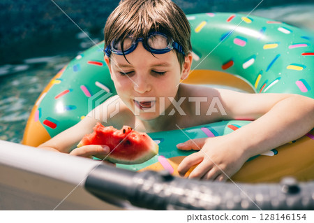 Boy in float with goggles enjoying juicy watermelon in backyard pool on sunny summer afternoon 128146154