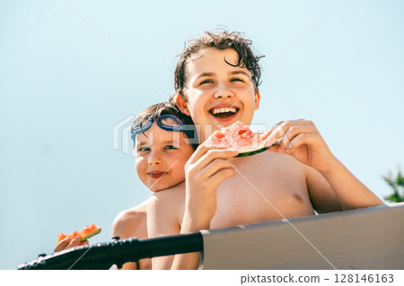Two cheerful boys eating watermelon together and enjoying a sunny summer day in the pool Two cheerful boys eating watermelon together and enjoying a sunny summer day in the pool 128146163