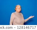 Caucasian woman in her 70s showing open palm offering new medical beauty procedure, anti-aging product, posing isolated over blue studio background, copy space for advertising text concept. 128146717