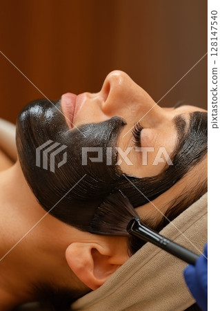 Woman relaxing with black charcoal face mask applied by brush during spa facial treatment 128147540